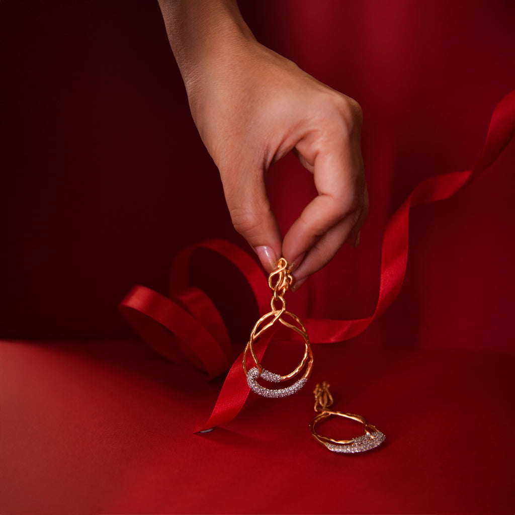 A hand displaying Backstage Earrings, a crystal-encrusted hoop earring with a second earring resting below, against a red backdrop